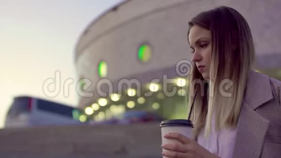 晚城的一个年轻女孩拿着咖啡和电话等着开会冻僵了扔在夹克上悲伤视频的预览图