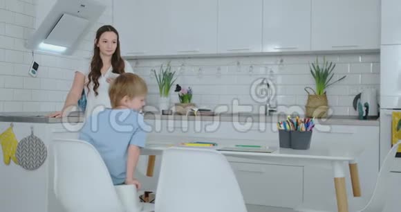 妈妈在厨房洗碗两个儿子坐在办公桌前用彩色铅笔在纸上画画年轻的家庭妈妈视频的预览图