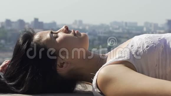 可爱的女人躺在屋顶边缘享受阳光城市天空线的背景视频的预览图