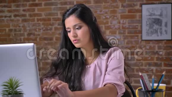 聪明漂亮的考恩女士正集中精力在她的笔记本电脑上打字而她正坐在她的桌面上的砖砌办公室里视频的预览图
