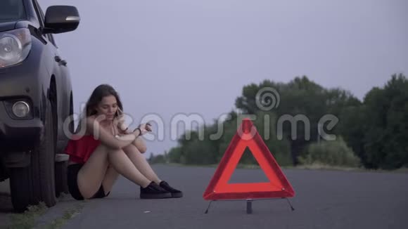 心烦意乱的女孩坐在她失事的车旁边年轻的女人坐在失事的车旁边悲伤的女人正在打电话视频的预览图