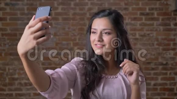 年轻快乐的黑发白种人女孩是塔丽克自拍与她的手机显示两个手指和微笑同时视频的预览图
