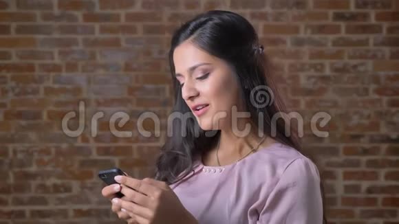 可爱的白种人女人正在滚动她的手机并在屏幕上有体贴的面部打字答案在砖块背景上视频的预览图