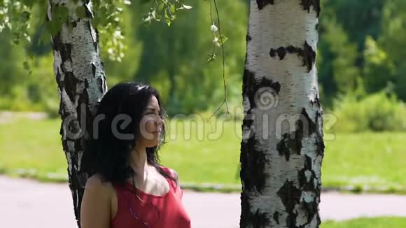 穿着红裙子的女人站在公园的桦树下等待约会伙伴视频的预览图
