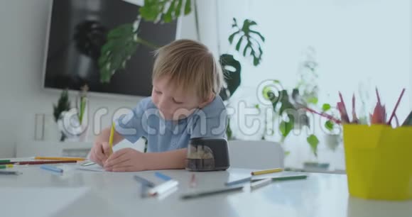 穿着蓝色衬衫的微笑男孩坐在客厅的桌子旁用铅笔在纸上画画视频的预览图