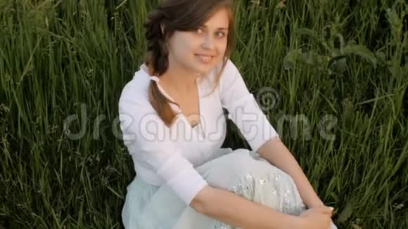 年轻美丽的农村女孩坐在草地上的田野里微笑着女人的情感和春天人类的美丽和概念视频的预览图