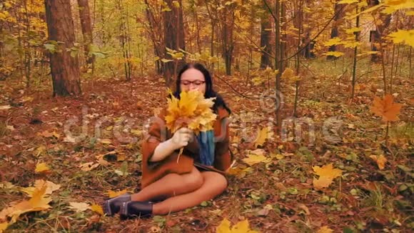 戴眼镜的年轻开朗的女人坐在秋天公园的草地上手里拿着枫叶视频的预览图