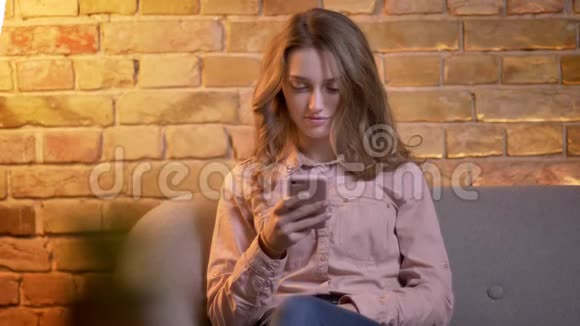 年轻迷人的少女坐在沙发上带着智能手机在舒适的家中微笑成镜头的画像视频的预览图