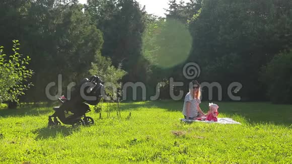 戴着眼镜戴着帽子的年轻漂亮妈妈和她的小女儿坐在绿色的草坪上公园里有树视频的预览图