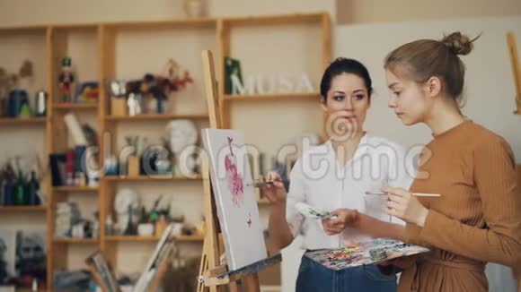 有经验的绘画老师正在和她的女学生交谈她站在画架上的照片附近讨论艺术品视频的预览图