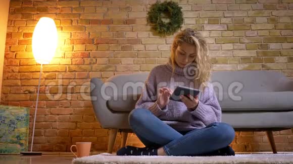 一位成年白种人金发女性在舒适的室内坐在地板上时使用平板电脑的特写镜头视频的预览图