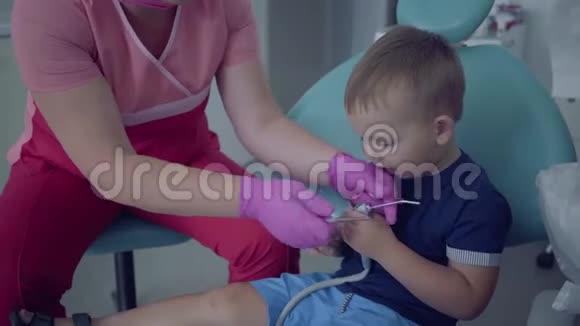 戴着口罩和手套的牙医准备检查坐在椅子上的无忧无虑的小男孩的牙齿视频的预览图