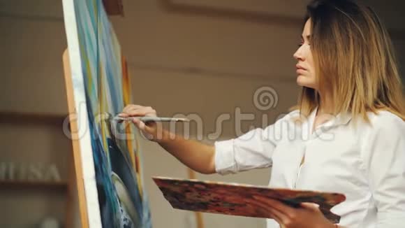 漂亮的年轻女子穿着白色衬衫在画布上用油画画笔和调色板绘制海景视频的预览图