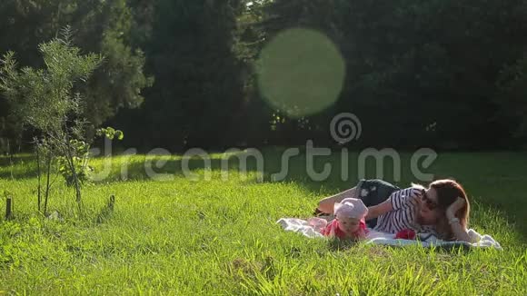 快乐的年轻美丽的母亲戴着眼镜她的小女儿躺在绿色的草坪上铺着白色的毯子视频的预览图