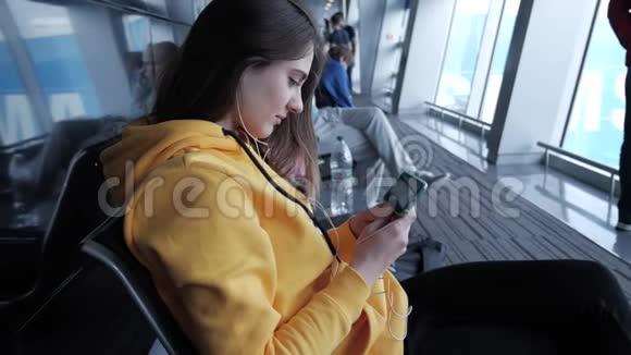 女人在机场等待登机花时间在智能手机上看电影戴着耳机的女孩盯着手机视频的预览图