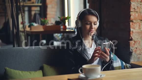 戴着耳机的年轻女子在现代咖啡馆里用智能手机听音乐视频的预览图