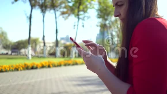 漂亮的商务女性穿着红色连衣裙在城里散步并在智能手机上使用应用程序视频的预览图