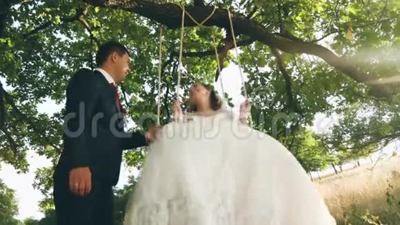 快乐的年轻夫妇新娘和新郎穿着婚纱在公园里荡秋千夏天在橡树枝上荡秋千视频的预览图