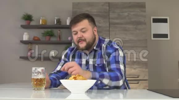一个长着胡子的男人穿着格子衬衫坐在厨房里喝着带薯片的啤酒吃饭的乐趣慢吞吞的视频的预览图
