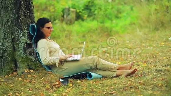 一个戴眼镜的女人赤脚坐在公园的树下用笔记本电脑工作视频的预览图