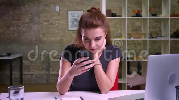 可爱的白种人商业女孩坐在室内在办公室里精确地看着屏幕她正在滚动她的智能手机视频的预览图
