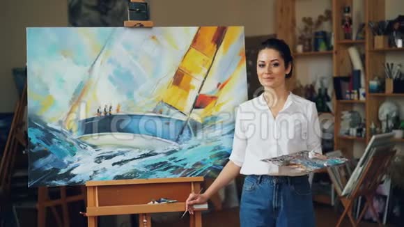 一幅美丽的年轻女画家站在现代画的旁边看着摄像机和微笑的画像视频的预览图