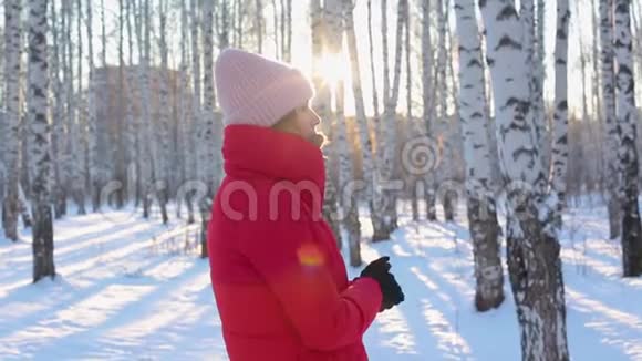 穿着红色冬衣的可爱的年轻女子站在画报城市公园里在寒冷的阳光明媚的日子里有桦树在期待中结冰视频的预览图