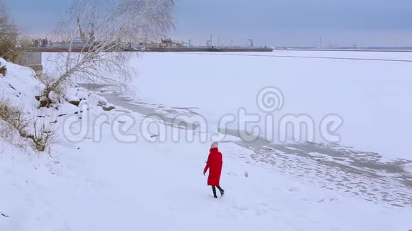 背面风景年轻女子穿着红色长夹克走在白雪覆盖的河岸上弯曲的生长着桦树视频的预览图