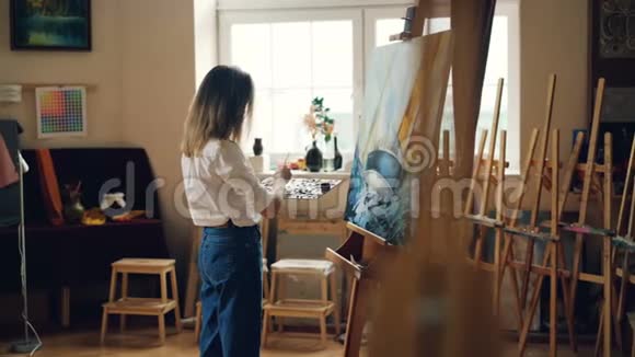 瘦削的金发女孩正拿着画笔用油画颜料作画描绘了在室内工作的海景视频的预览图