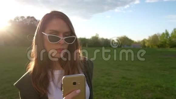 美丽的年轻女人在公园里用金色智能手机自拍视频的预览图