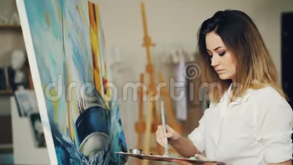 漂亮的年轻女士正忙着在车间里用明亮的油漆和彩色调色板作画女孩穿着休闲装视频的预览图