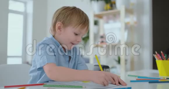 穿着蓝色衬衫的微笑男孩坐在客厅的桌子旁用铅笔在纸上画画视频的预览图