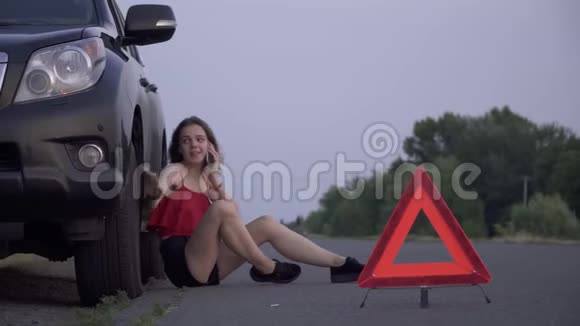 心烦意乱的女孩坐在她失事的车旁边年轻的女人坐在失事的车旁边悲伤的女人正在打电话视频的预览图