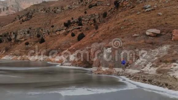 在史诗般的岩石背景下一位女摄影师沿着冰冻的山湖岸边漫步视频的预览图