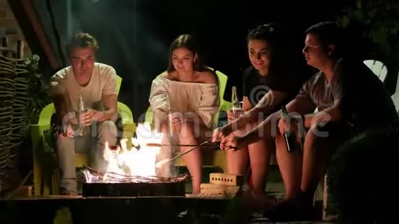 快乐的朋友晚上坐在篝火旁烤香肠聊天喝啤酒视频的预览图