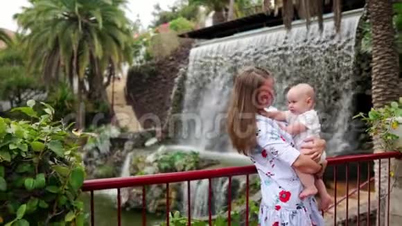 年轻的妈妈抱着一个孩子和儿子一起玩在公园的瀑布附近微笑和欢笑同时去旅行视频的预览图