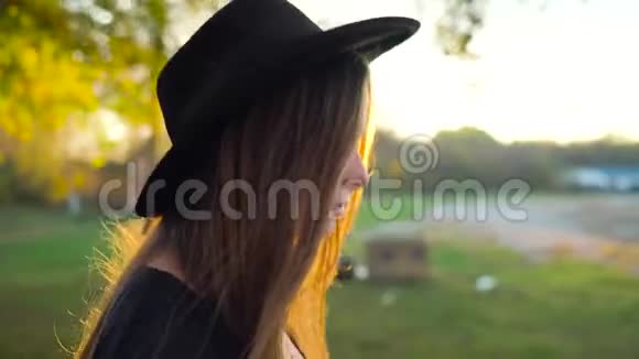 美丽的微笑女孩戴着黑色的帽子背景是日落走在户外慢动作视频的预览图