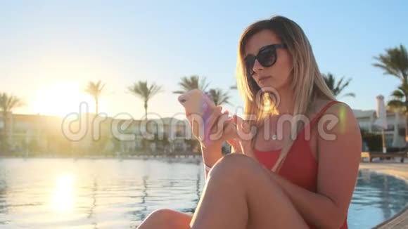 迷人的年轻女孩穿着红色泳衣在游泳池附近的智能手机上交谈的肖像太阳下的比基尼女孩视频的预览图