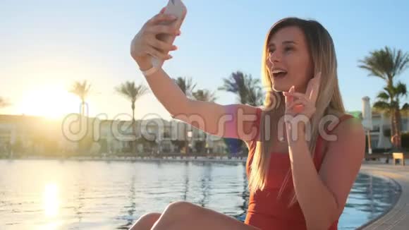 一张漂亮的金发美女的肖像红色泳衣正在游泳池附近放松用智能手机自拍视频的预览图