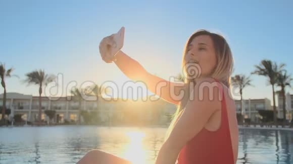 一张漂亮的金发美女的肖像红色泳衣正在游泳池附近放松用智能手机自拍视频的预览图