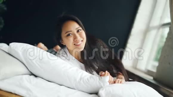 穿着白色T恤的快乐亚洲女士在床上翻滚的肖像微笑着看着镜头表达积极的情绪视频的预览图