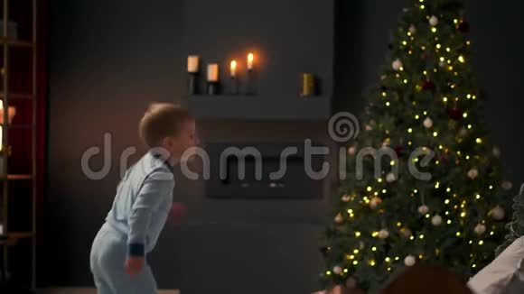 快乐的小男孩跳上床在圣诞树的背景上扔一个枕头视频的预览图