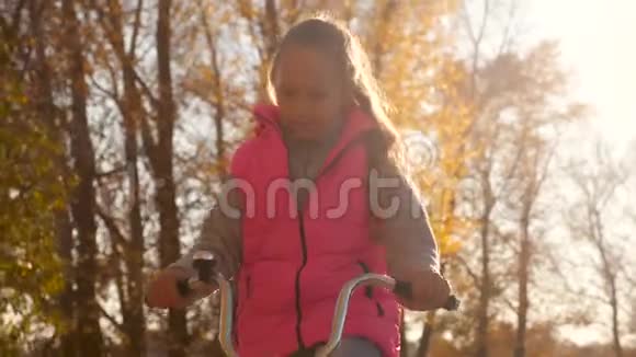 穿着粉红色夹克的快乐女孩骑着自行车在秋天公园的黄树背景下视频的预览图