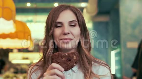 一个迷人的女孩坐在咖啡馆里吃着饼干看着相机视频的预览图