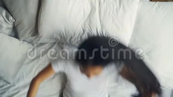 穿着白色T恤的漂亮的亚裔深色头发的女孩躺在舒适的床上铺着白色亚麻布摸着她的头发视频的预览图
