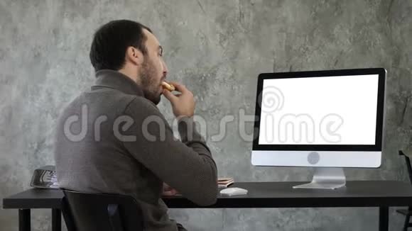 年轻人一边吃东西一边在办公室里看电脑上的东西白色显示器视频的预览图