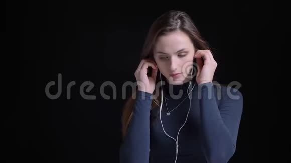 年轻苗条的白种人女孩在耳机里听音乐在黑色背景下跳舞的肖像视频的预览图