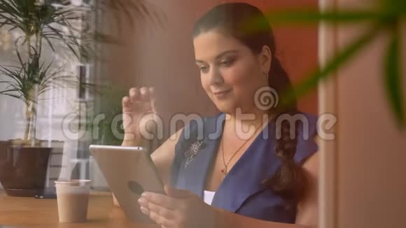 肥胖的白种人可爱的女孩拿着她的杯子在咖啡馆的窗户附近的设备上游泳视频的预览图