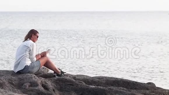 一个年轻的女人正拿着智能手机坐在海边的岩石海岸上视频的预览图