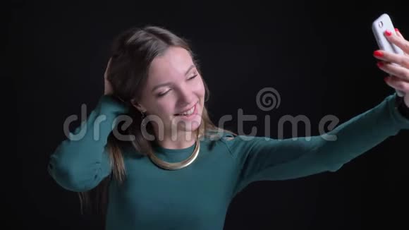 年轻的黑发白种人女孩在黑色背景下微笑地使用智能手机制作自拍照片的肖像视频的预览图